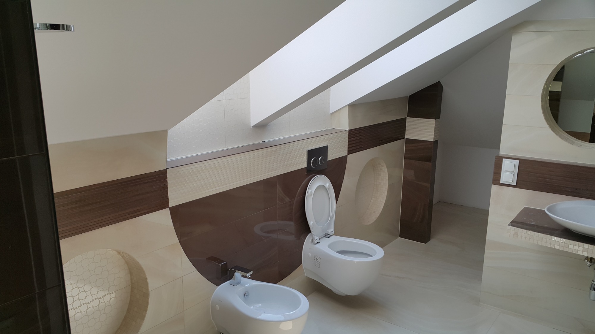 Nowoczesna łazienka na poddaszu z toaletą podwieszaną, bidetem i okrągłym lustrem, wykończona płytkami w odcieniach beżu i brązu z dekoracyjnymi okręgami.