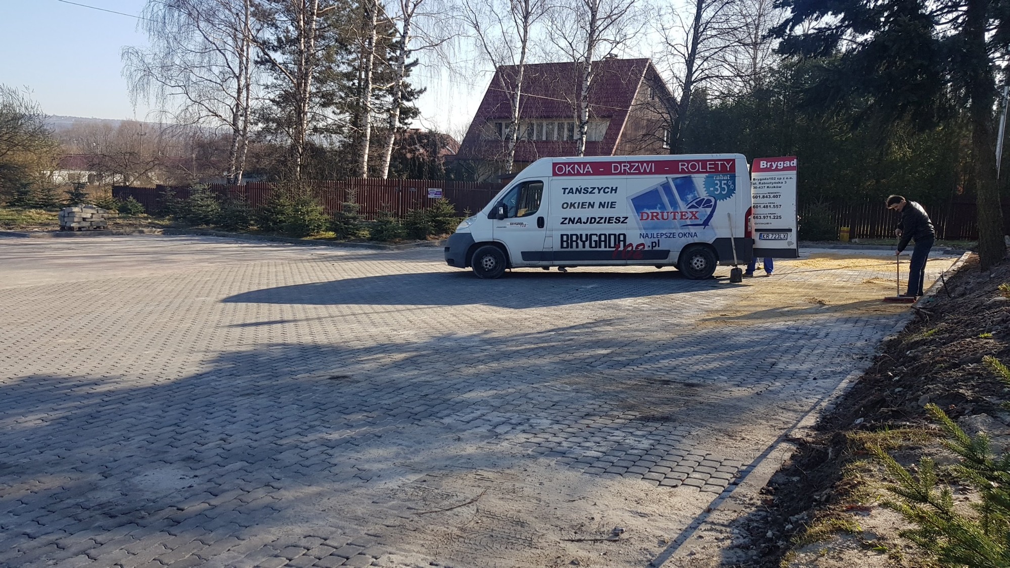 Brygada, biały samochód dostawczy z reklamą okien DRUTEX, pracownik zamiata resztki materiału na parkingu z kostki brukowej, w tle budynek z czerwonym dachem i drzewa bez liści.