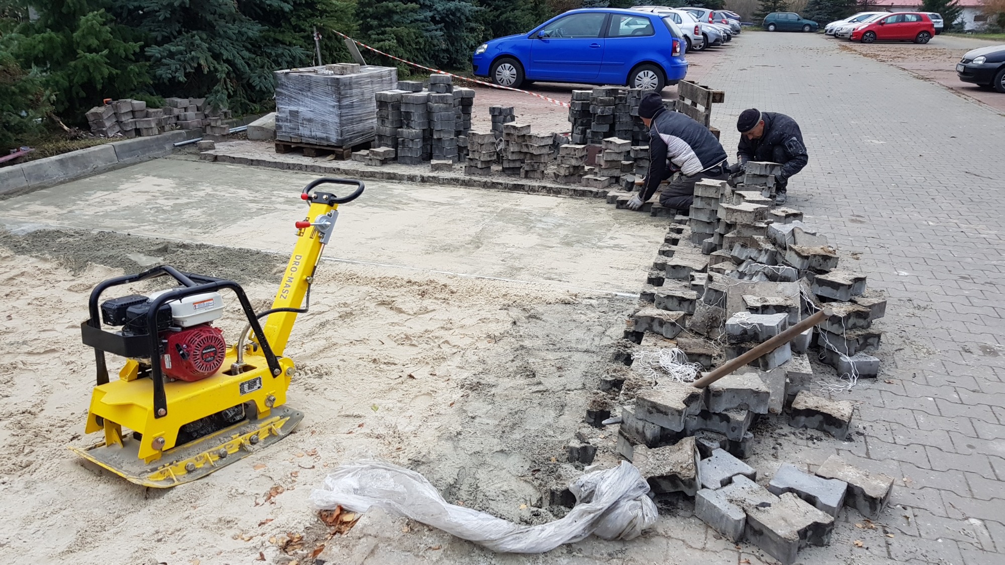Dwóch robotników układa szarą kostkę brukową na piaskowym podłożu, żółta zagęszczarka obok, w tle zaparkowane samochody i drzewa.