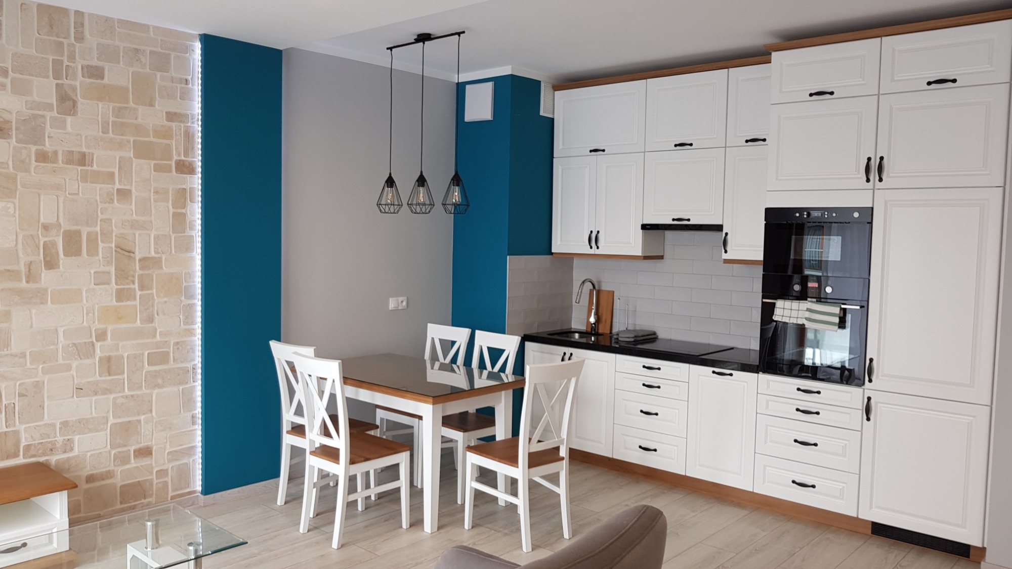 Jasna kuchnia z białymi szafkami i drewnianymi akcentami, czarnym blatem i sprzętami AGD, stół z krzesłami w stylu skandynawskim, ściana z dekoracyjną cegłą i turkusowym pasem, oświetlenie wiszące...