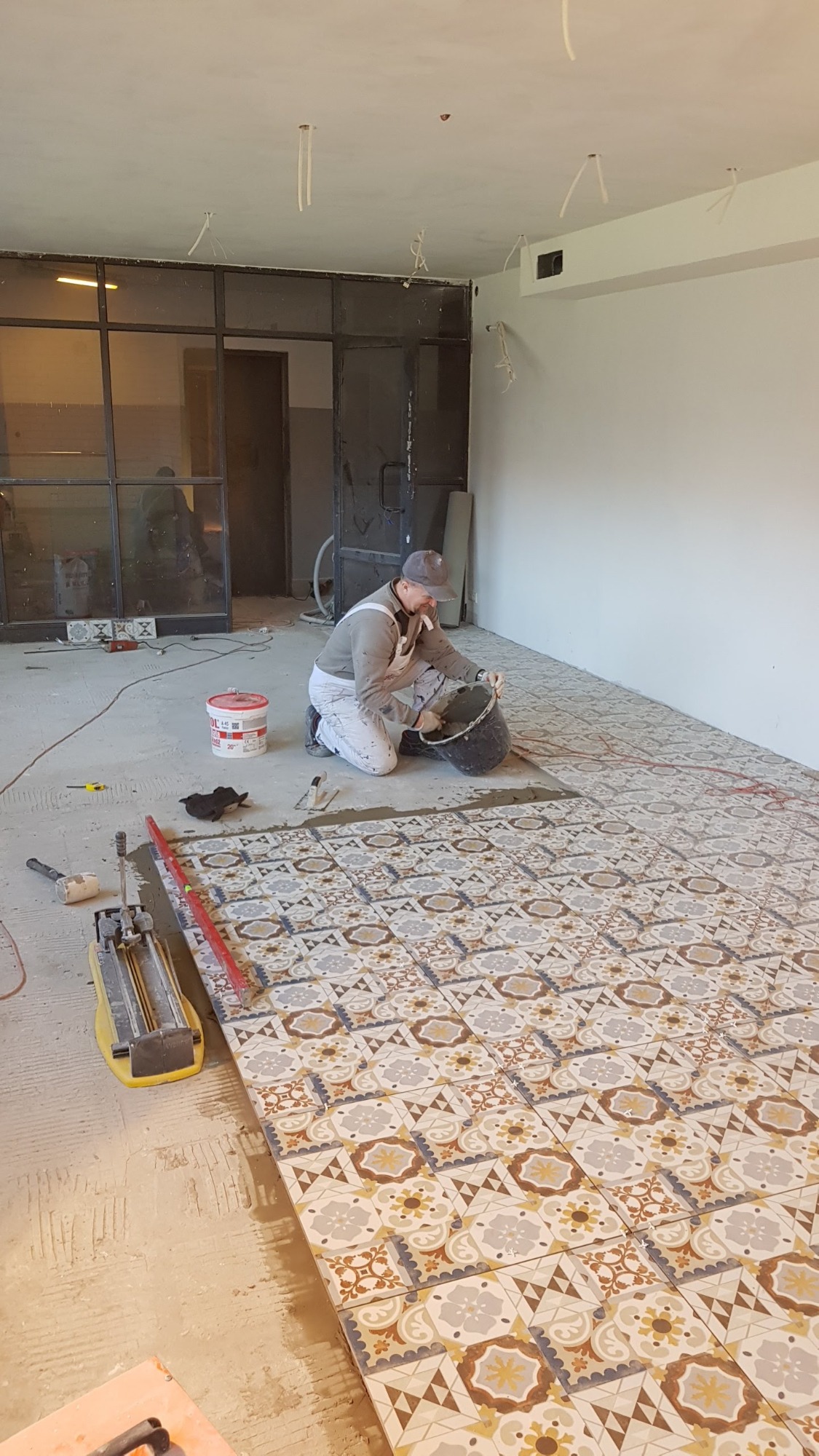Układanie wzorzystych płytek podłogowych w trakcie remontu, widoczny glazurnik na kolanach z wiadrem zaprawy, narzędzia glazurnicze i częściowo ułożona podłoga.