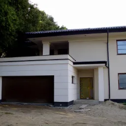 Dwukondygnacyjny dom z jasną elewacją i brązową bramą garażową, drzwi wejściowe zabezpieczone folią ochronną, widoczne elementy wykończeniowe.