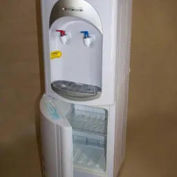 Biały dystrybutor wody z otwartą szafką chłodzącą na naczynia, widoczne krany z oznaczeniem ciepłej i zimnej wody.