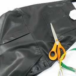 Fragment czarnych, skórzanych spodni roboczych na szelkach w Komorowie, częściowo uszytych, z nożyczkami z pomarańczowymi uchwytami i miarką krawiecką obok, leżących na białym tle z widocznym...