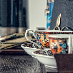 Stylowe filiżanki w orientalny wzór na ciemnym, drewnianym biurku w biurze. W tle czajnik elektryczny i inne akcesoria biurowe. Przytulna aranżacja przestrzeni biurowej.