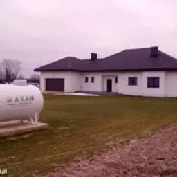 Biały zbiornik na gaz płynny marki AXAN posadowiony na betonowych bloczkach przed nowoczesnym, parterowym domem z ciemnym dachem.