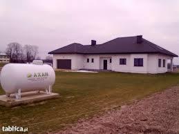 Biały zbiornik na gaz płynny marki AXAN posadowiony na betonowych bloczkach przed nowoczesnym, parterowym domem z ciemnym dachem.