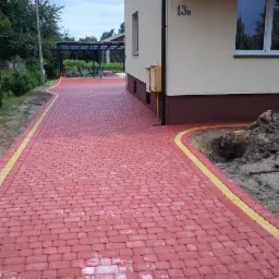 Czerwona kostka brukowa z żółtym obramowaniem tworzy ścieżkę przy budynku 13B. W tle widoczny zadaszony taras i zieleń. Wykopana ziemia po prawej stronie.