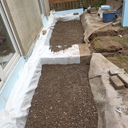 Jacek Bińczycki JACK BAU - Fundament domu z izolacją: styropian, biała folia i żwir. Widoczne okno i fragment ściany budynku. Prace w toku, teren wokół częściowo wykopany.