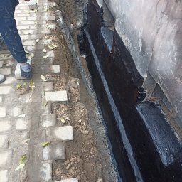 Jacek Bińczycki JACK BAU - Prace przy izolacji fundamentów: robotnik układa kostkę brukową wzdłuż ściany z nałożoną czarną izolacją. Widoczny fragment wykopu i użyte narzędzia.