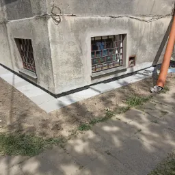 Fragment budynku z oknami piwnicznymi po izolacji fundamentów i ułożeniu chodnika z kostki. Widoczna rura spustowa i fragment chodnika.