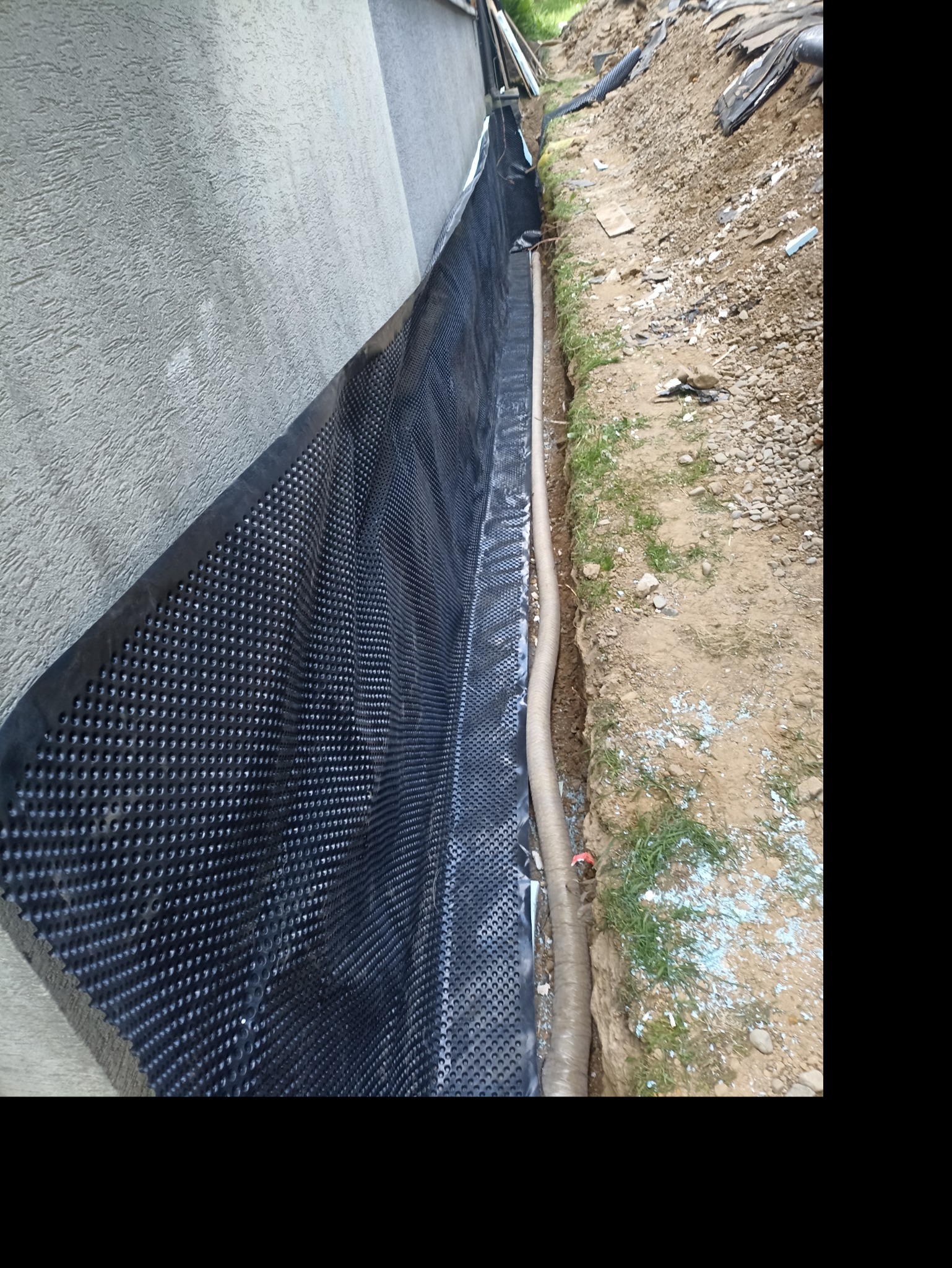 Czarna folia izolacyjna z wypustkami na fundamencie, rura drenażowa w wykopie. Widoczne ślady białego proszku. Prace ziemne w toku.