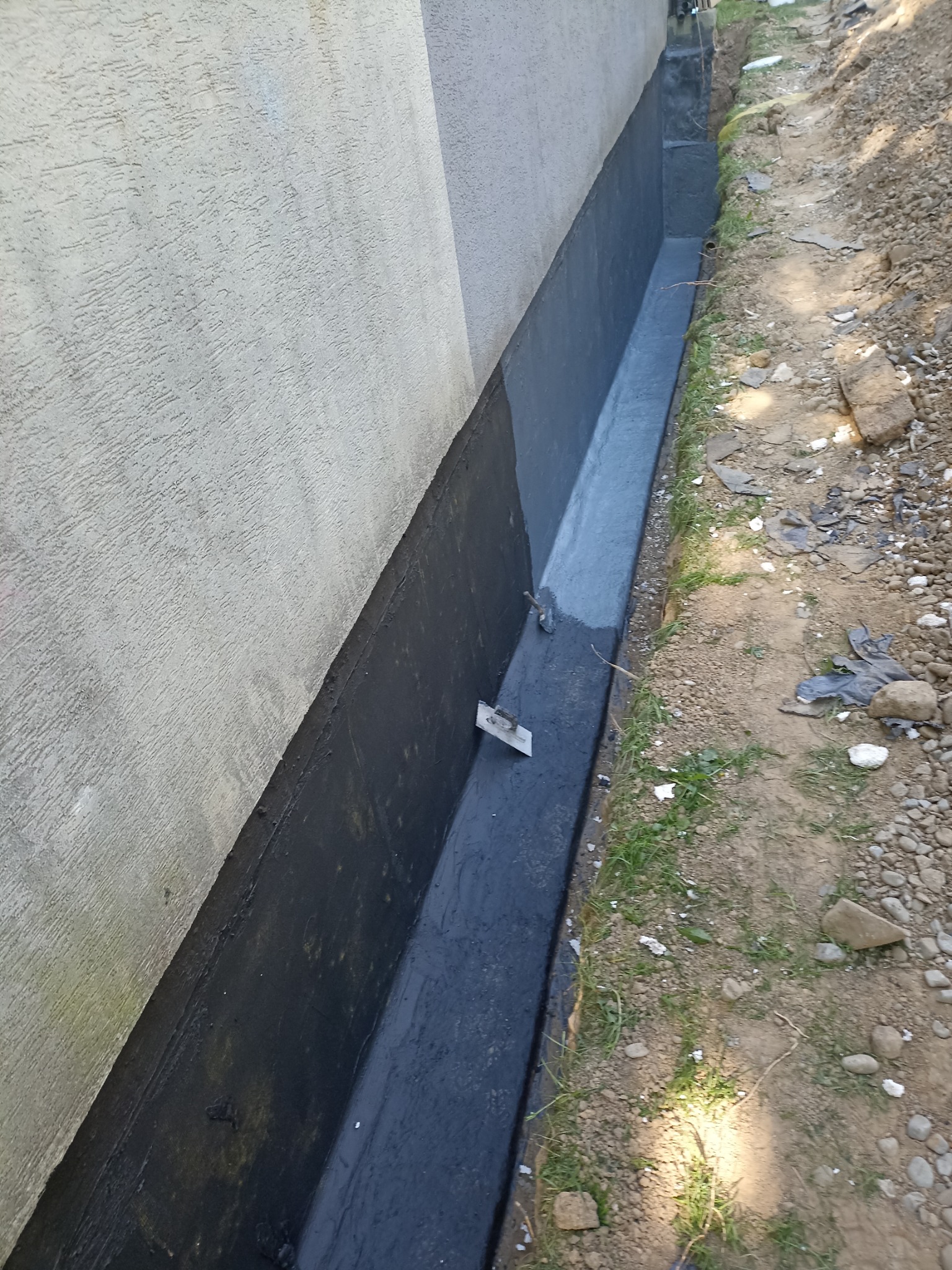 Świeżo zaizolowany fundament budynku z czarną hydroizolacją, widoczna kielnia murarska, grunt wokół. Prace ziemne przy budynku.