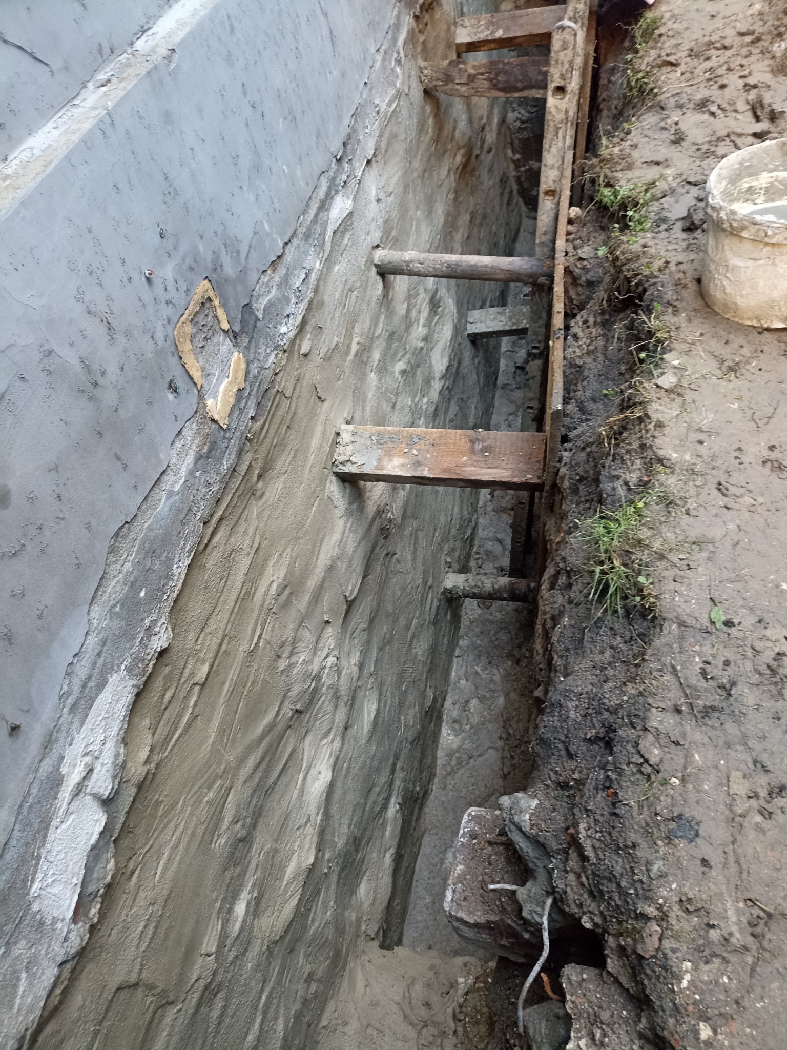 Zabezpieczony wykop fundamentowy z drewnianą konstrukcją wsporczą i świeżo nałożoną warstwą izolacji pionowej, widoczne ślady napraw na ścianie.