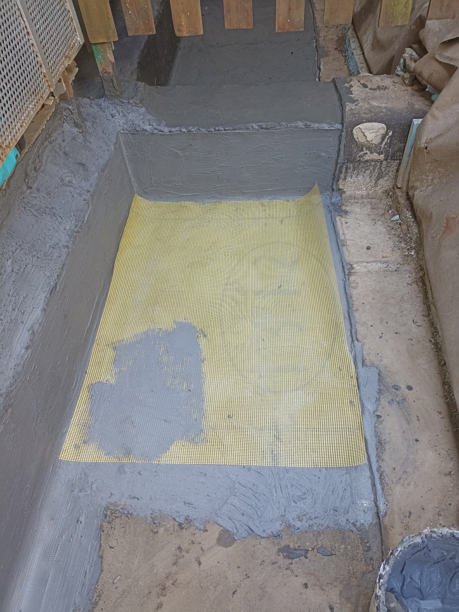 Żółta siatka na podłożu, częściowo pokryta szarą zaprawą, otoczona świeżo nałożoną szarą izolacją. Widoczne drewniane elementy konstrukcyjne.