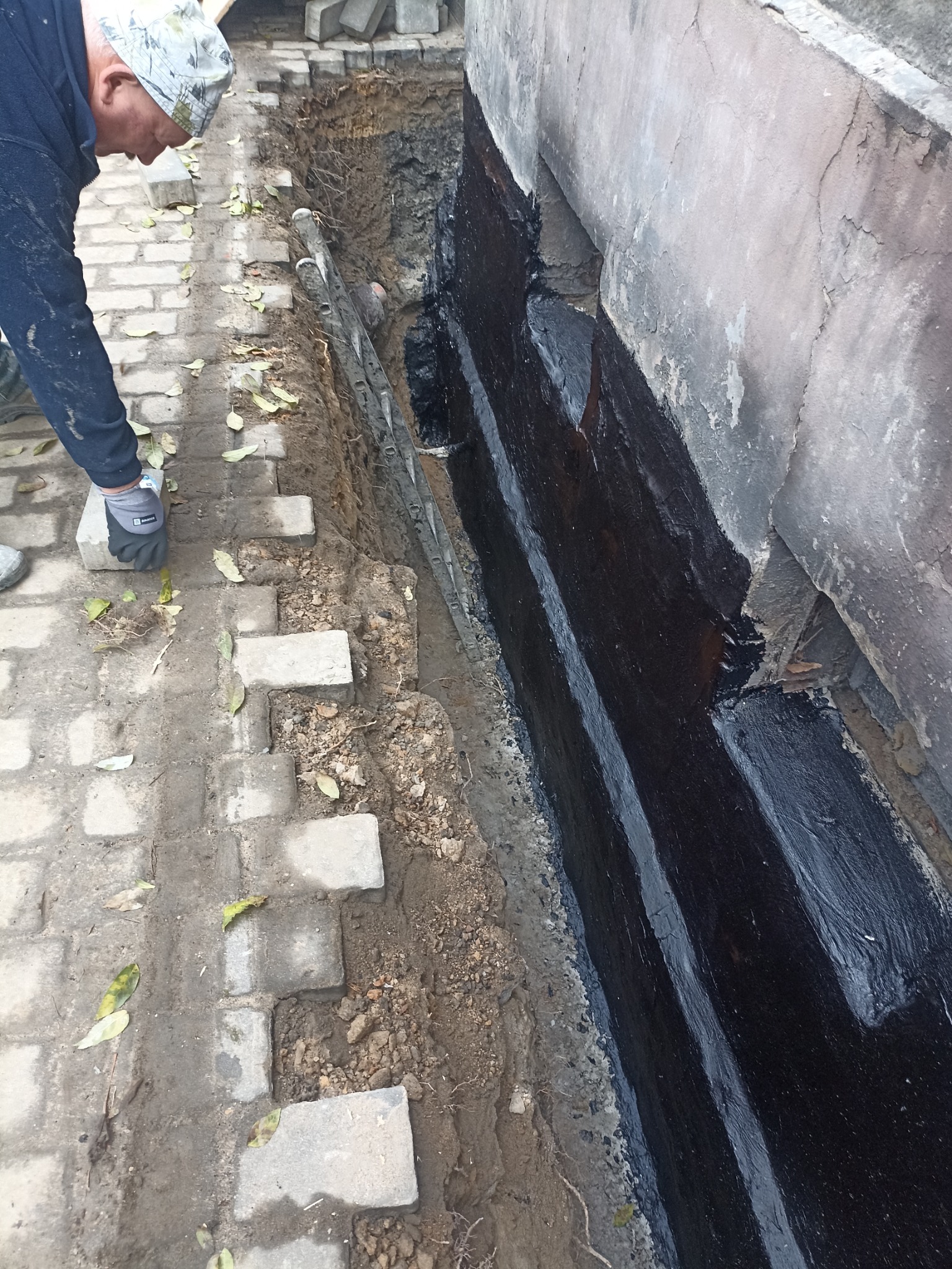 Prace przy izolacji fundamentów: robotnik układa kostkę brukową wzdłuż ściany z nałożoną czarną izolacją. Widoczny fragment wykopu i użyte narzędzia.