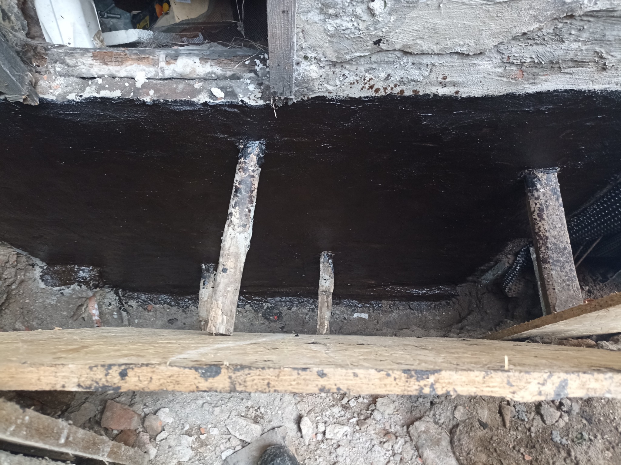 Izolacja fundamentów z użyciem czarnej, gęstej substancji. Widoczne drewniane podpory i fragmenty ściany. Prace izolacyjne w toku, fundament zabezpieczony przed wilgocią.