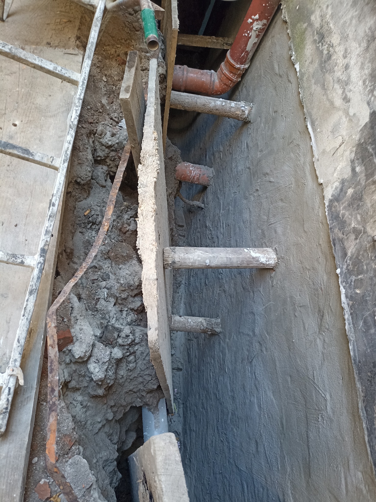 Świeżo otynkowana ściana fundamentowa z wystającymi drewnianymi kołkami, obok wykop z widocznymi fragmentami rur i deskowaniem. Detal prac izolacyjnych.