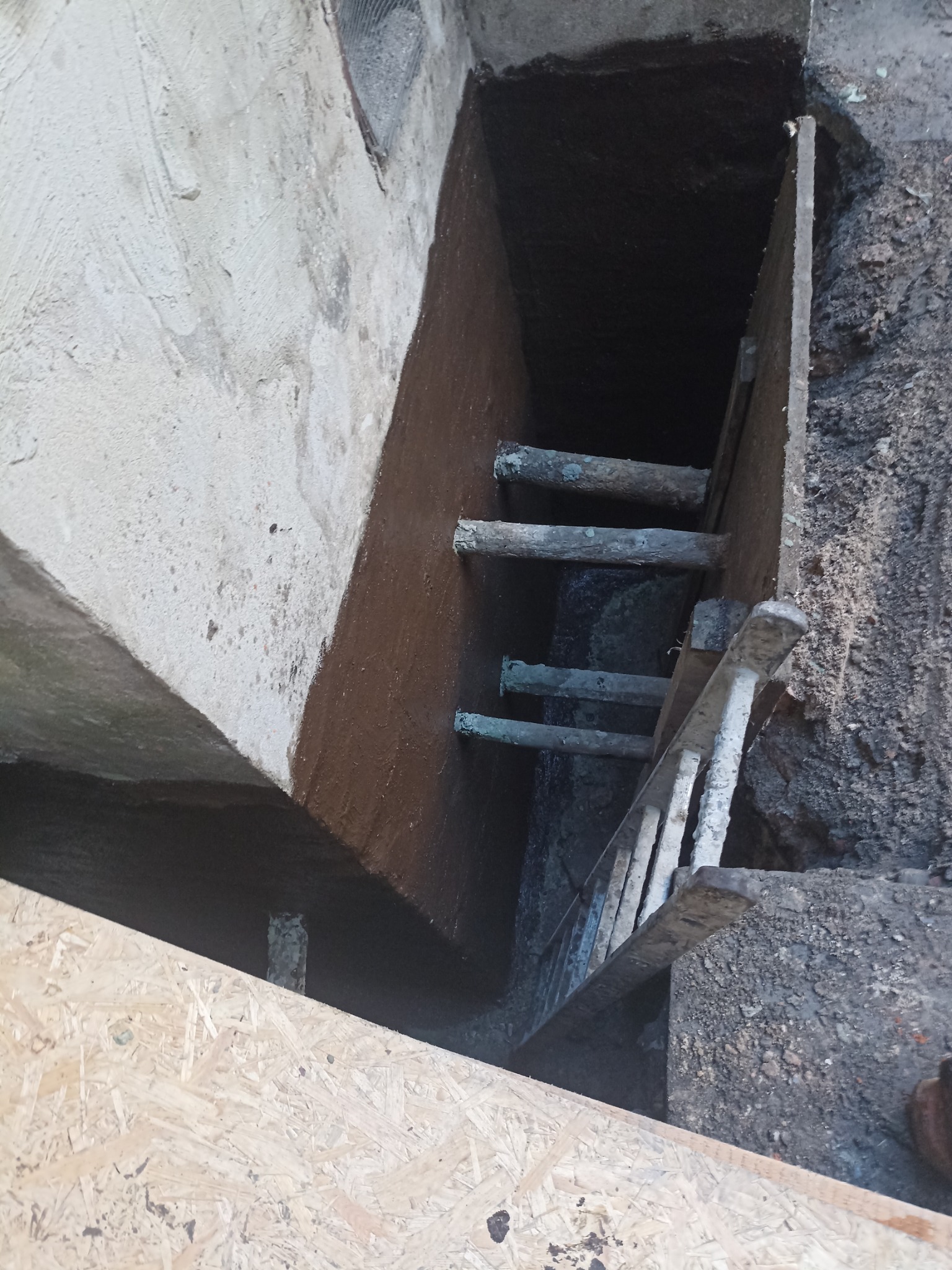 Zabezpieczone wykopalisko fundamentowe z izolacją pionową, widoczne drewniane szalunki i drabina. Grunt wokół wykopu. Ujęcie z góry.