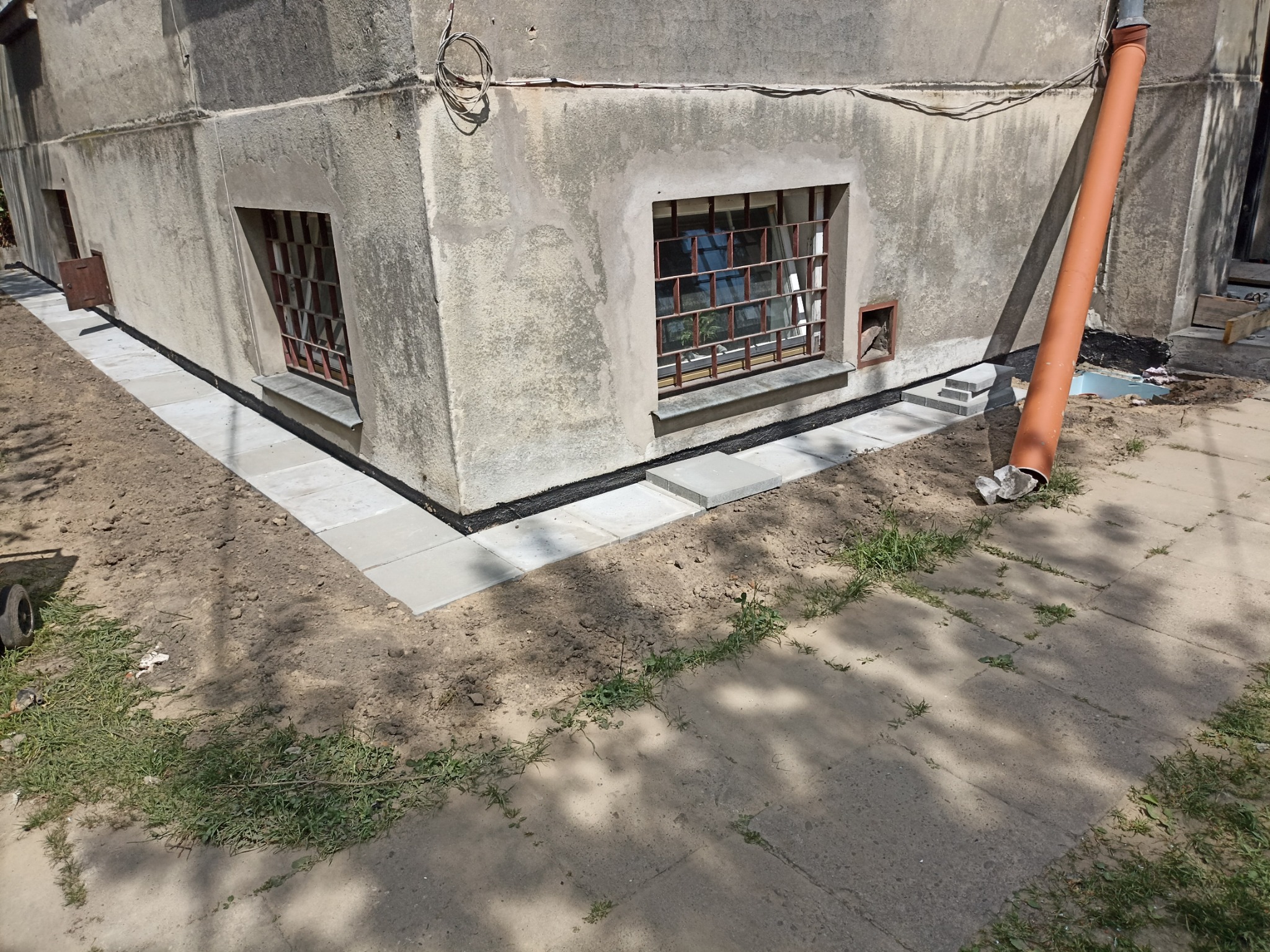 Fragment budynku z oknami piwnicznymi po izolacji fundamentów i ułożeniu chodnika z kostki. Widoczna rura spustowa i fragment chodnika.
