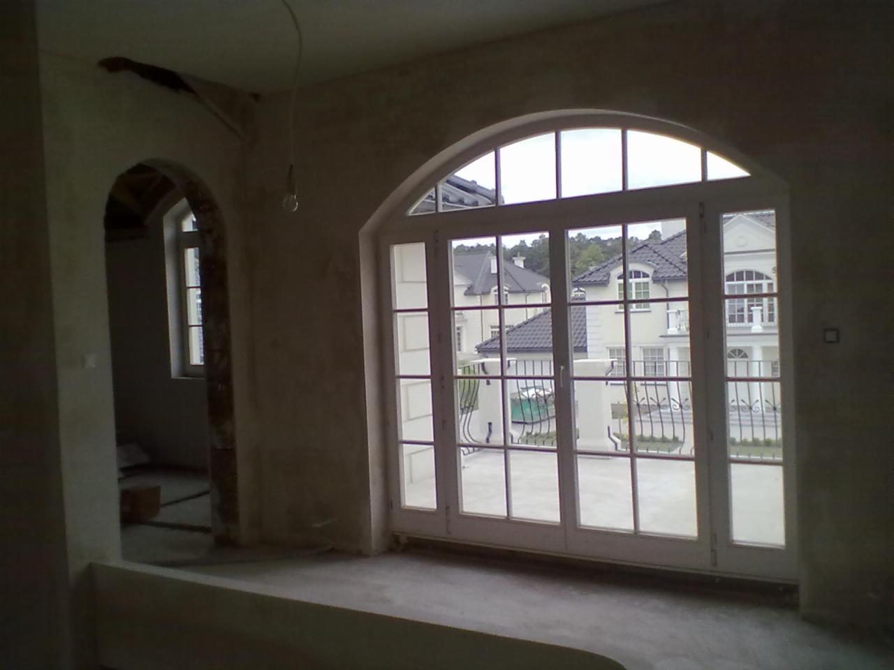 Wnętrze w trakcie prac tynkarskich: surowe ściany z widoczną strukturą, duże okno łukowe z widokiem na zabudowę willową, surowy stan wykończenia.