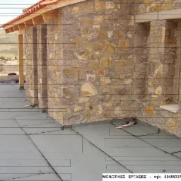 Budynek z kamienia z nowym pokryciem dachu, widok z góry na płaski dach z widocznymi łączeniami i elementami mocującymi. W tle wzgórza.