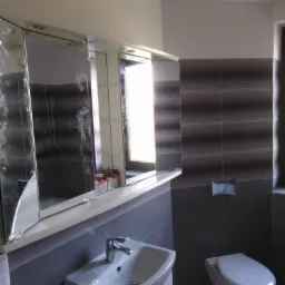 Umywalka z szafką, lustro z szafkami nad umywalką oraz toaleta w szaro-białej łazience z poziomymi pasami na ścianie.