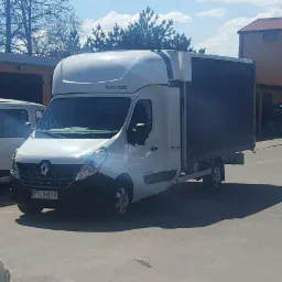 Biały Renault Master z plandeką i nadbudówką typu Twin Cab, zaparkowany na placu w słoneczny dzień, widoczny numer rejestracyjny PTU 9401A.