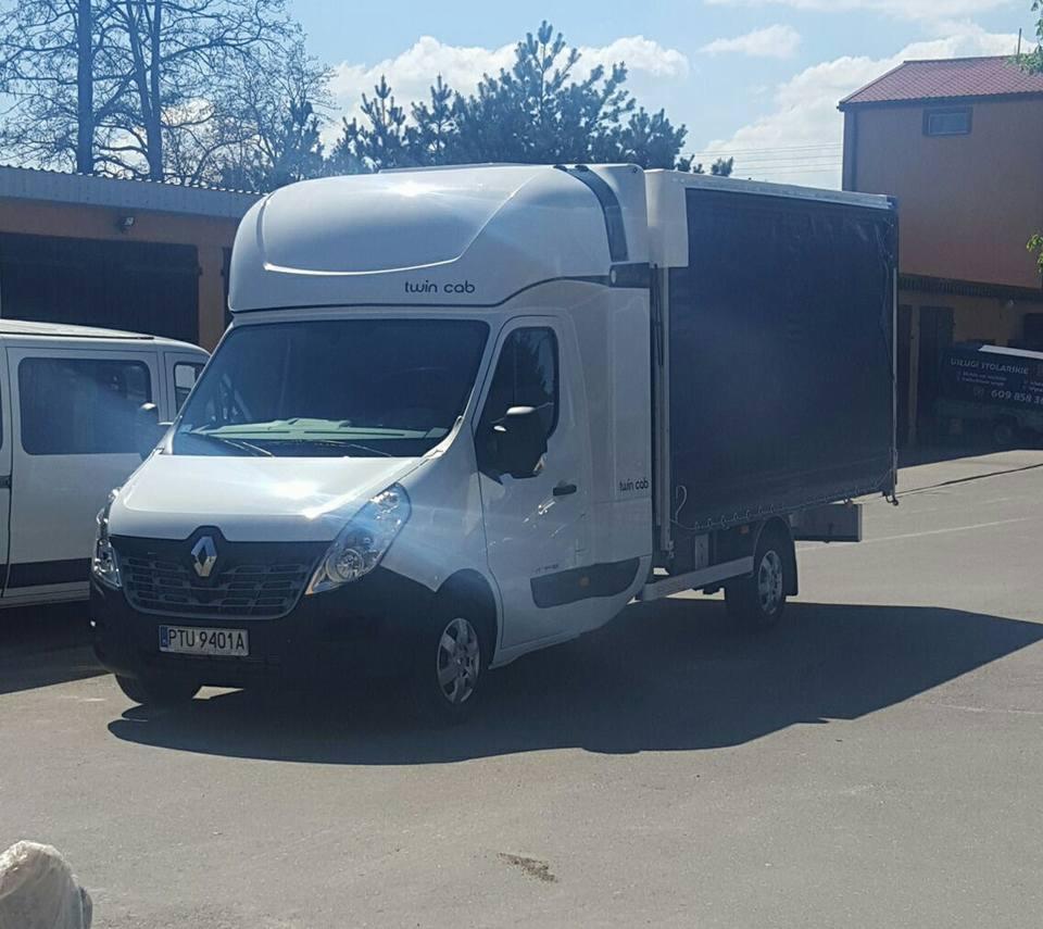 Biały Renault Master z plandeką i nadbudówką typu Twin Cab, zaparkowany na placu w słoneczny dzień, widoczny numer rejestracyjny PTU 9401A.