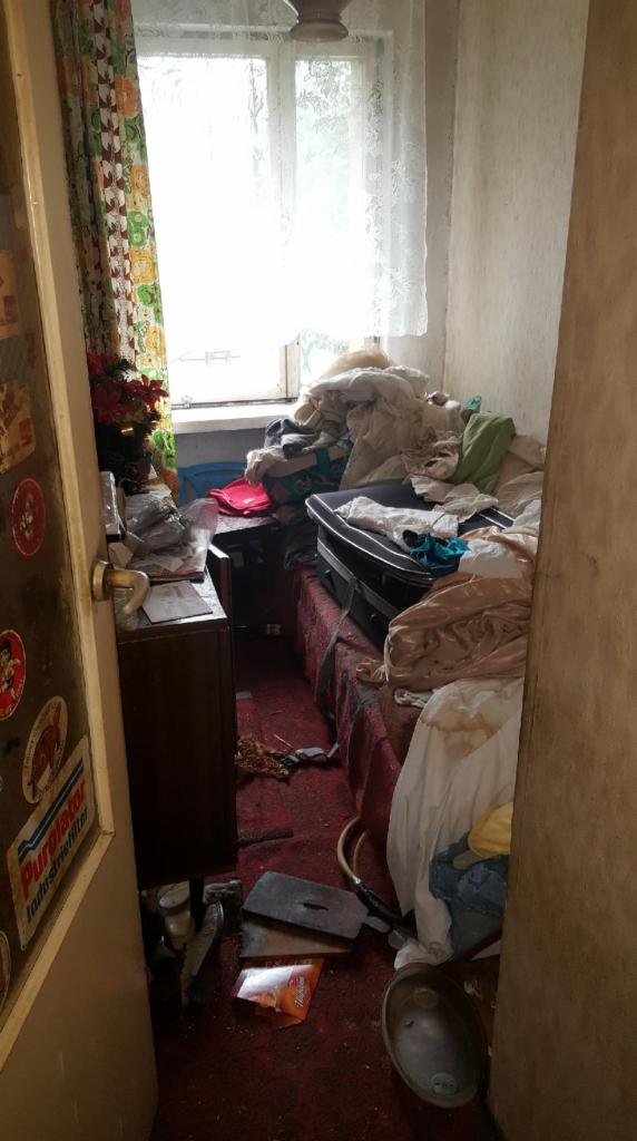 Zaniedbane wnętrze małego pokoju z łóżkiem zawalonym rzeczami, widoczne narzędzia i śmieci na podłodze, przez otwarte drzwi widać naklejki na szafce.