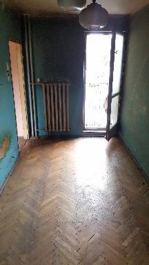 Pusty pokój przed remontem z parkietem ułożonym w jodełkę, stary grzejnik żeberkowy, turkusowe ściany i otwarte drzwi balkonowe w bloku.