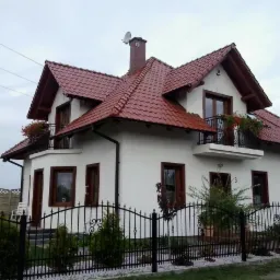 Biały dom jednorodzinny z czerwoną dachówką, balkonami z kwiatami i czarnym kutym ogrodzeniem, widok z zewnątrz w pochmurny dzień.