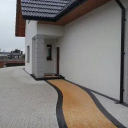 Nowoczesny, jednopiętrowy dom z jasną elewacją, ciemnym dachem i wybrukowanym podjazdem z dekoracyjnym, żółtym pasem prowadzącym do wejścia.