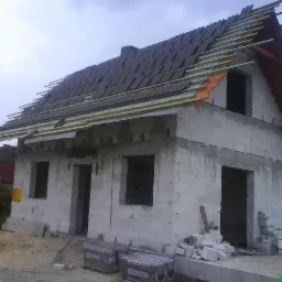 Wznoszony dwukondygnacyjny budynek z białych bloczków, częściowo pokryty dachówką ceramiczną Braas, widoczne przygotowane łaty dachowe i pustaki na placu budowy.