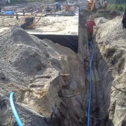 Wykop fundamentowy z zabezpieczeniem folią, widoczny pracownik w wykopie i sprzęt budowlany na placu budowy.