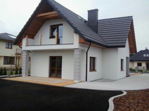 Nowoczesny dom murowany z białymi ścianami, ciemną dachówką i kominem, z balkonem z drewnianymi elementami, widok z zewnątrz na wykończony podjazd i ogród.