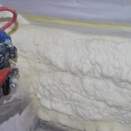 Aplikacja pianki PUR na ścianie za pomocą pistoletu natryskowego; widoczne rękawice ochronne i przewody zasilające.