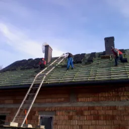Ekipa dekarzy na dachu domu jednorodzinnego podczas układania dachówki. Widoczne kominy i drabina oparta o niezakończoną elewację.