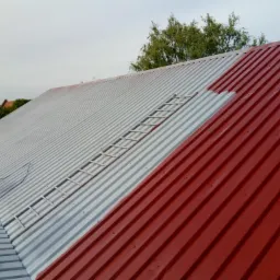 Częściowo pomalowany blaszany dach na czerwono, z widoczną drabiną i agregatem malarskim w tle.