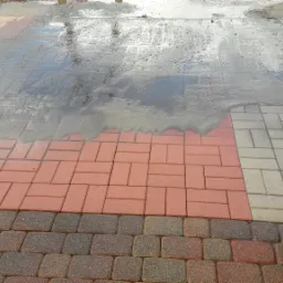 Mokry fragment chodnika z kostki brukowej w trzech odcieniach, z odbiciem nieba w kałuży i narożnikiem ceglanego muru