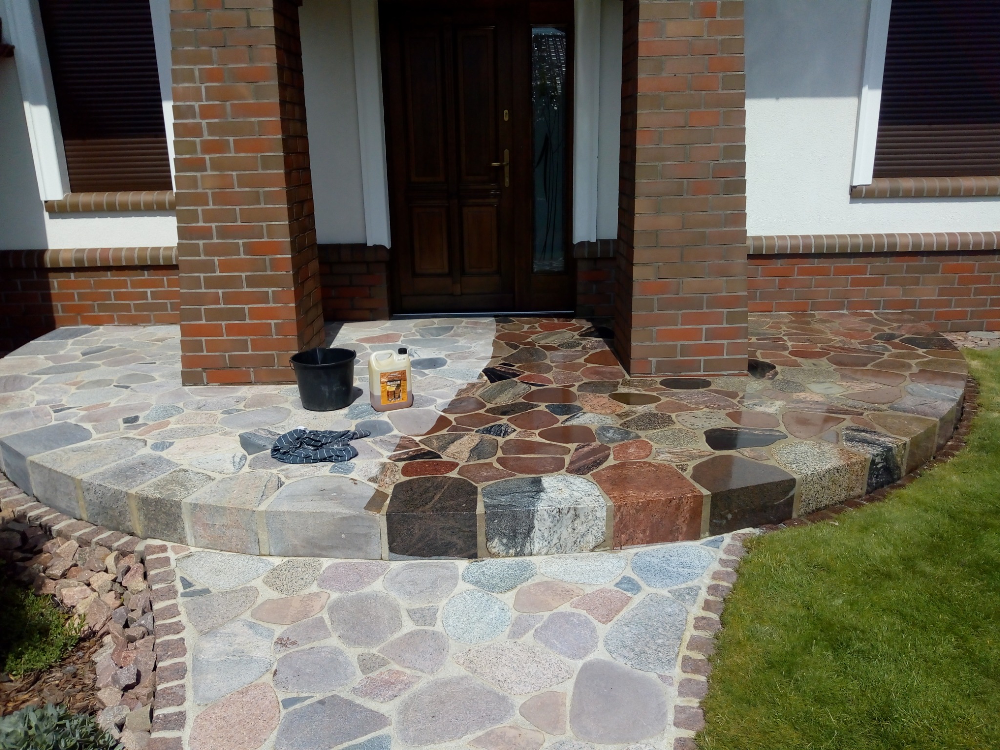 Kamienny podjazd przed domem w trakcie czyszczenia, widoczna różnica między umytą a nieumytą powierzchnią, wiadro z wodą i płyn do czyszczenia na boku.