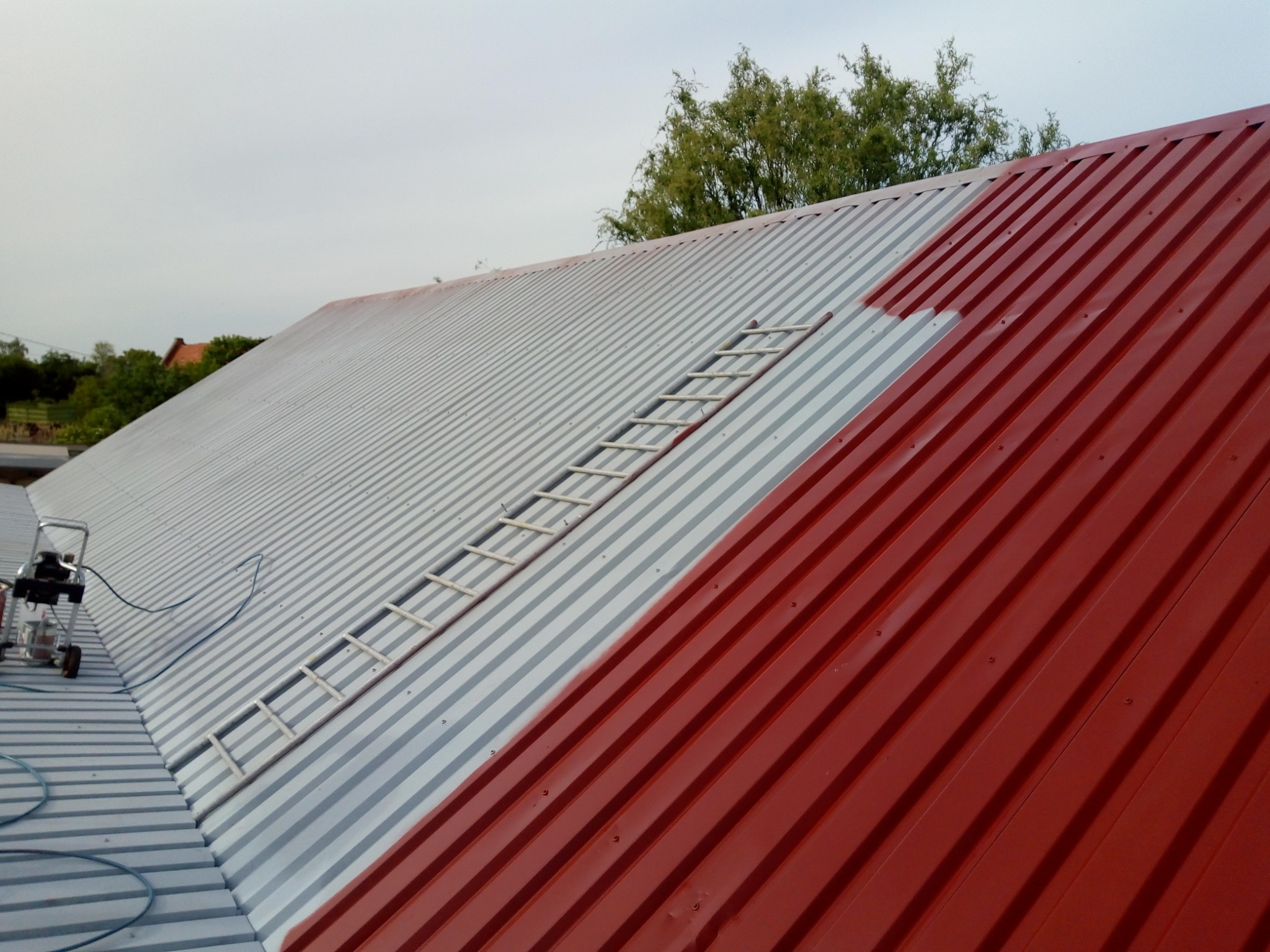 Częściowo pomalowany blaszany dach na czerwono, z widoczną drabiną i agregatem malarskim w tle.