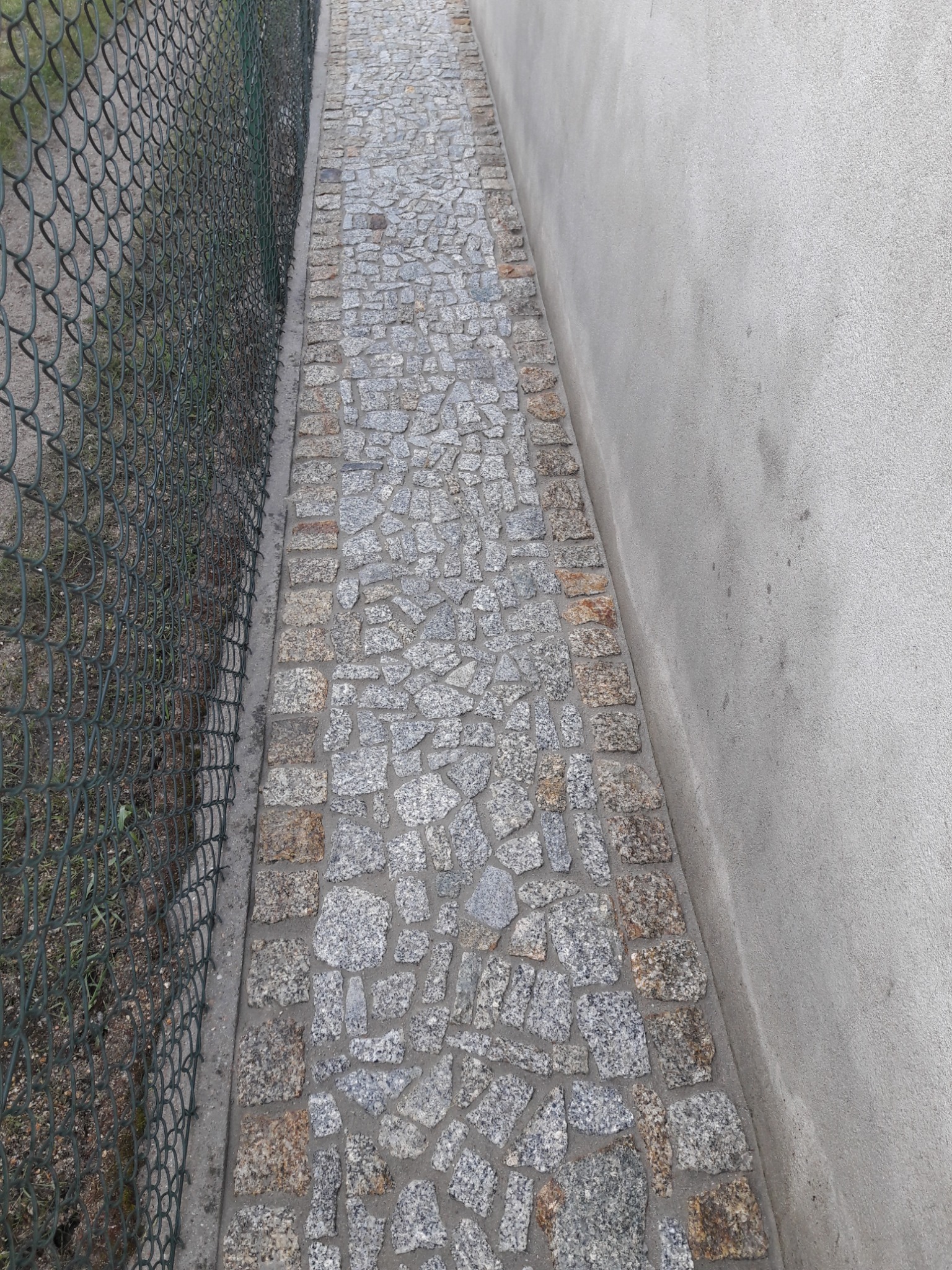 Granitowa ścieżka z obrzeżami z kostki brukowej, przylegająca do ogrodzenia z siatki i szarej ściany budynku.