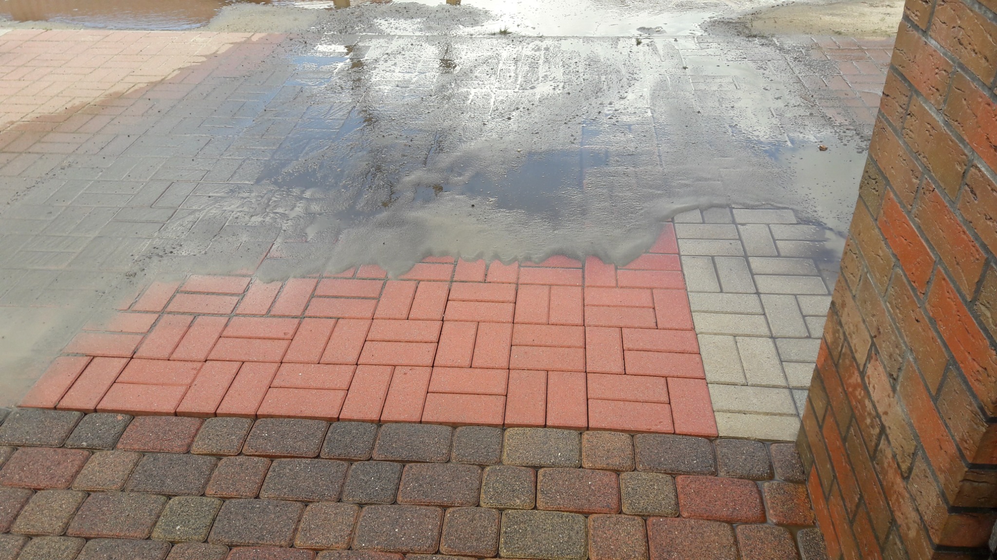 Mokry fragment chodnika z kostki brukowej w trzech odcieniach, z odbiciem nieba w kałuży i narożnikiem ceglanego muru