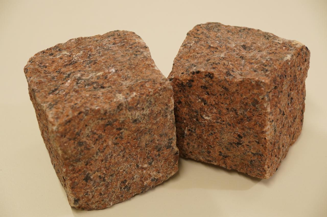 Dwie kostki granitowe w odcieniach czerwieni i brązu, o szorstkiej fakturze, na neutralnym tle. Widoczne nieregularne wzory i drobne ubytki w strukturze kamienia.