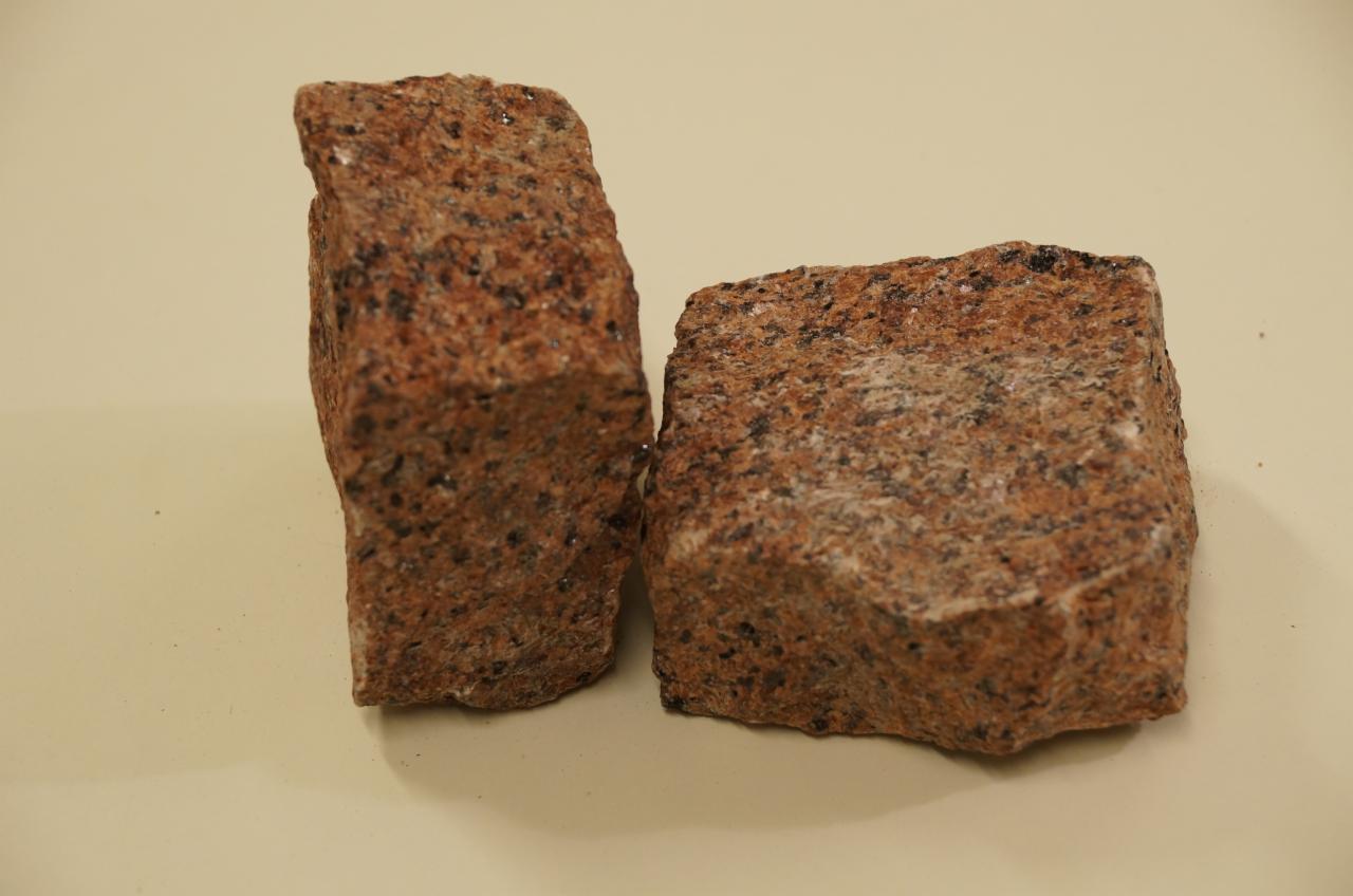 Dwie kostki granitowe w odcieniach czerwieni i szarości, o nieregularnych kształtach, ustawione obok siebie na gładkim, beżowym tle, eksponujące teksturę kamienia.