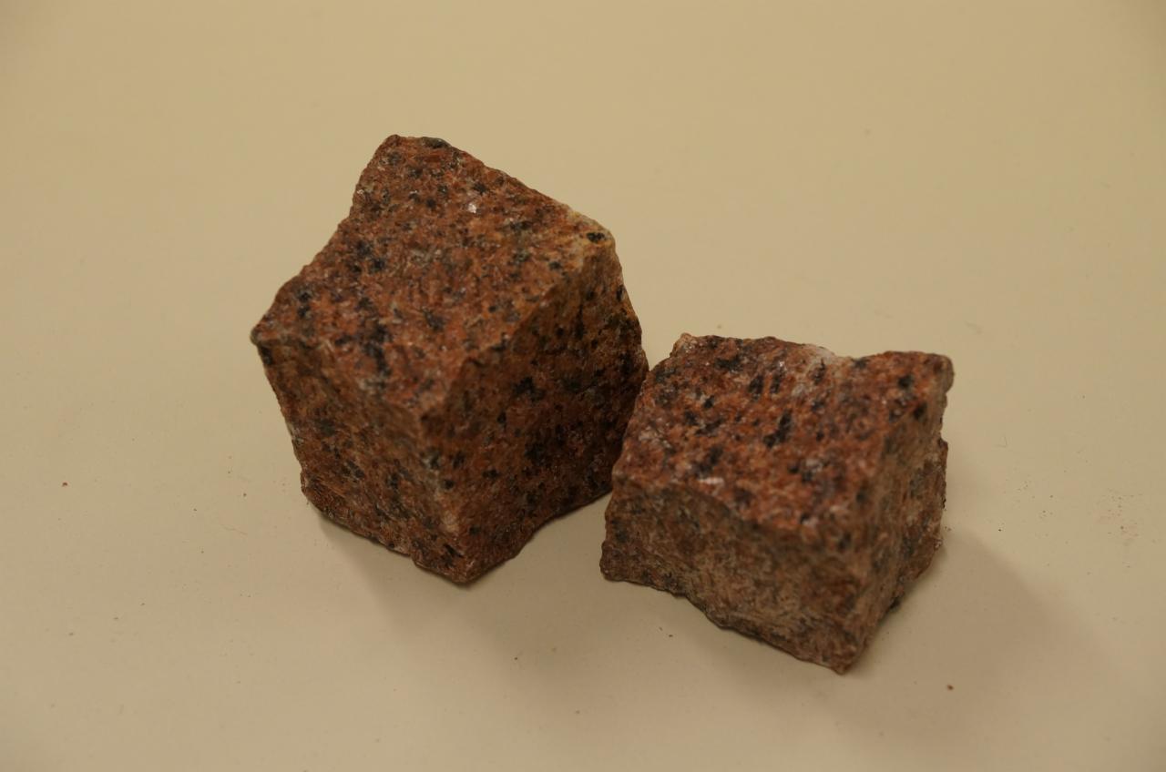 Dwa bloki kostki granitowej o nieregularnych kształtach, w odcieniach czerwieni i szarości, leżące na gładkim, beżowym tle. Widoczne drobne ziarna minerałów.