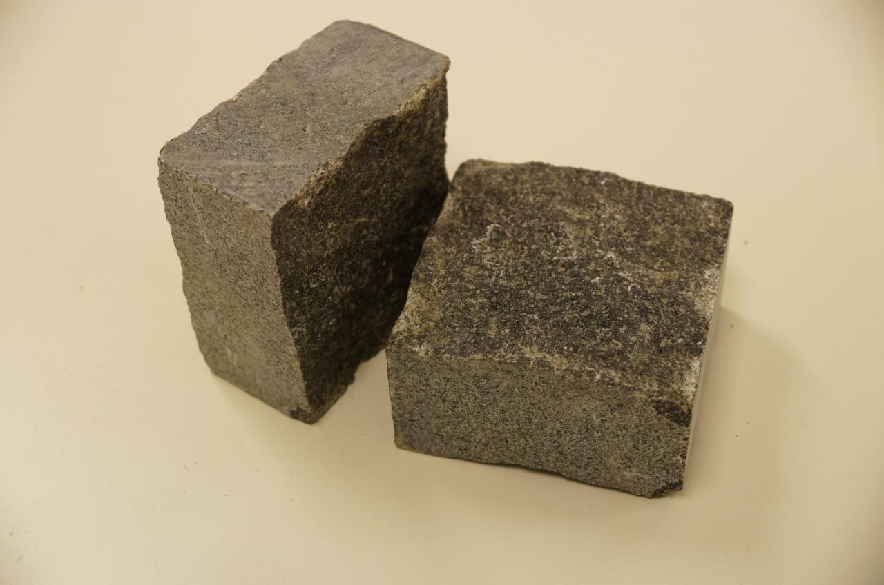 Dwie szare kostki granitowe o szorstkiej fakturze, jedna ustawiona pionowo, druga leżąca na beżowym tle, prezentacja materiału budowlanego.