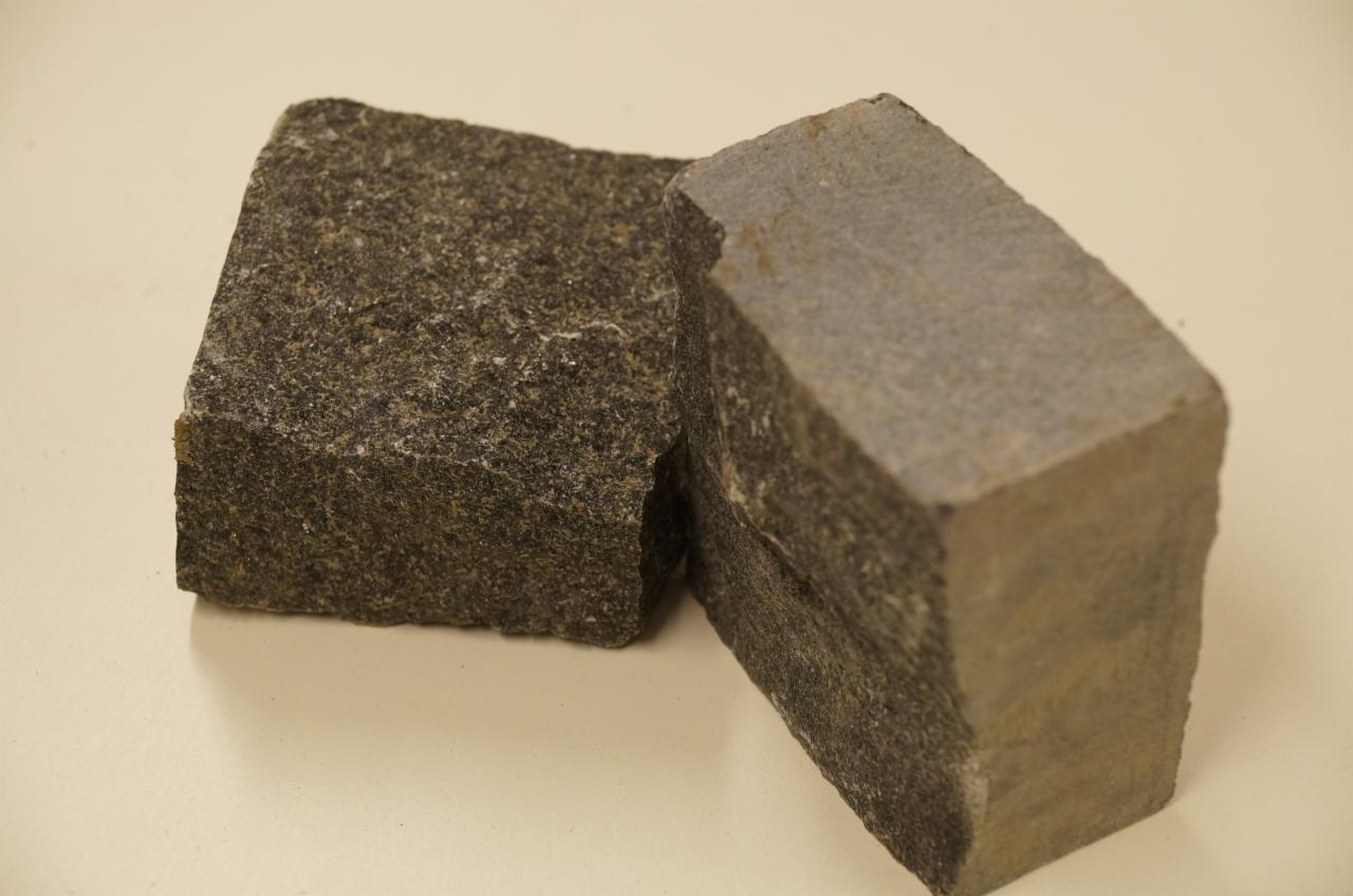 Dwie kostki granitowe o szorstkiej fakturze i nieregularnych krawędziach, w odcieniach szarości i brązu, leżące na jasnym, jednolitym tle. Bliskie ujęcie eksponuje detale kamienia.