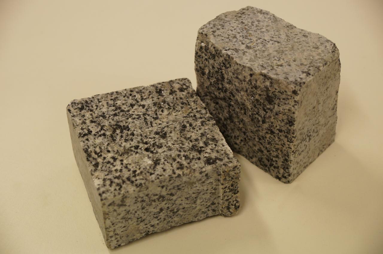 Dwa szare, surowe bloki z granitu o nieregularnej fakturze na jasnym tle. Jeden blok jest mniejszy i znajduje się bliżej, drugi jest większy i ustawiony z tyłu.